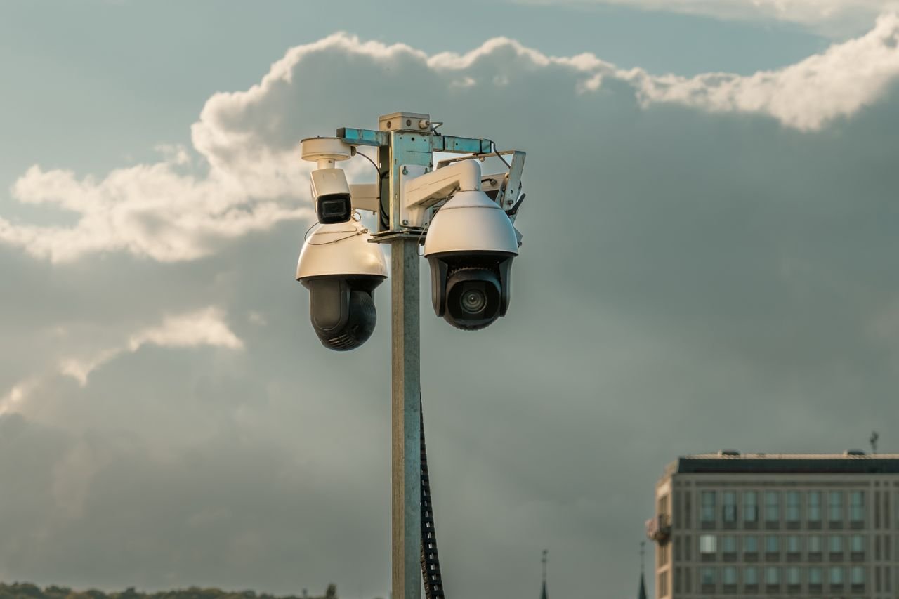 why-choose-us Two modern surveillance cameras mounted on a pole in an urban setting under a cloudy sky.
